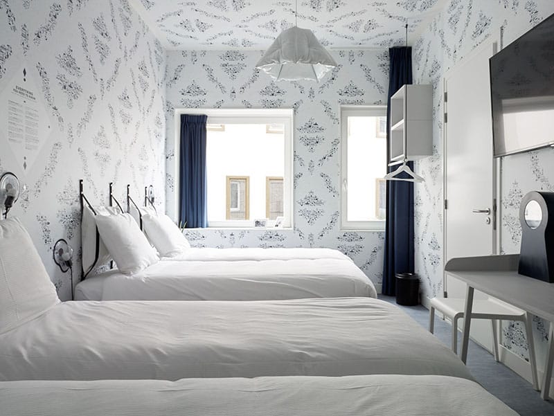 Helles Zimmer mit vier weißen Betten, zwei Fenstern mit blauen Vorhängen und einem Schreibtisch an der Wand | Kaboom-Hotel