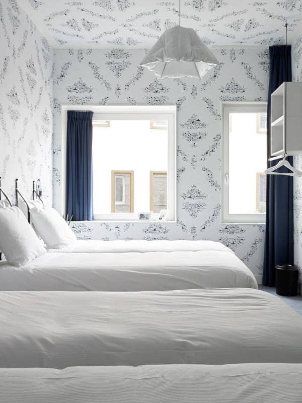 Lichte kamer met vier witte bedden, twee ramen met blauwe gordijnen en een bureau tegen de muur | Kaboom Hotel