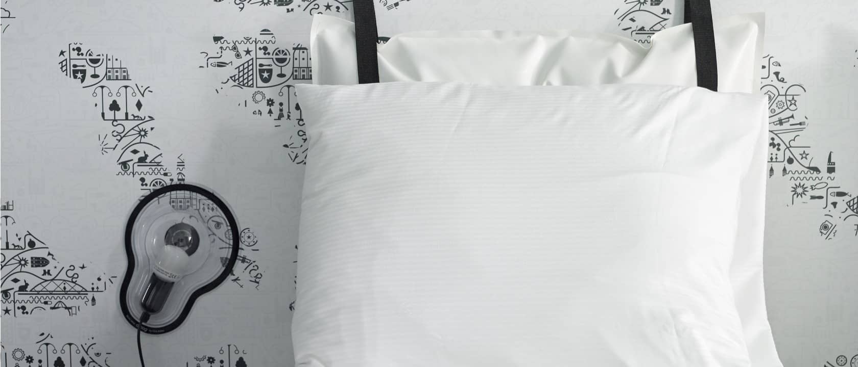 Overlappende kussens aan het hoofdeinde van het bed met een lamp tegen de muur ernaast | Kaboom Hotel