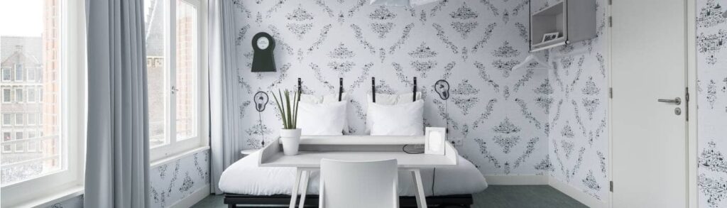 Zimmer mit zwei Fenstern, Doppelbett mit Schreibtisch davor und weißer Tür | Kaboom-Hotel